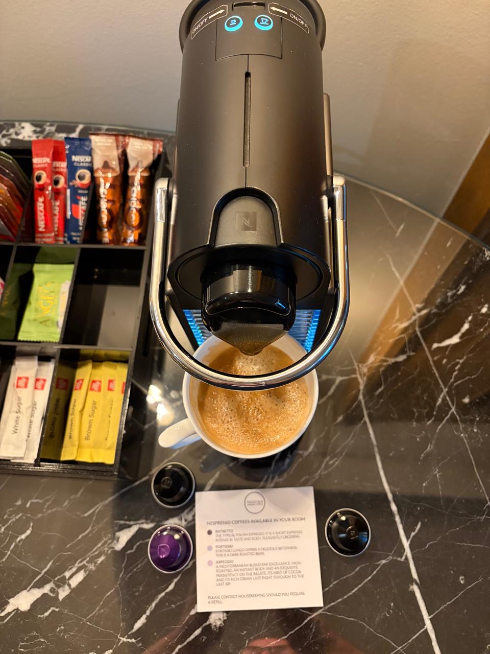 Top-down view of a Nespresso machine brewing espresso with golden crema, capsules flanking the cup on a marble counter