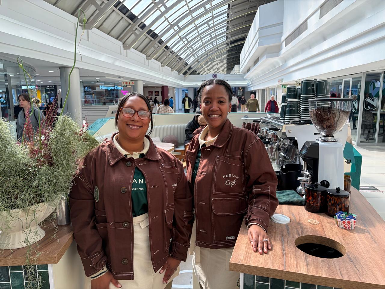 Two SOZO Foundation barista graduates smiling in their brown Fabiani Café jackets beside the espresso machine and hanging plants