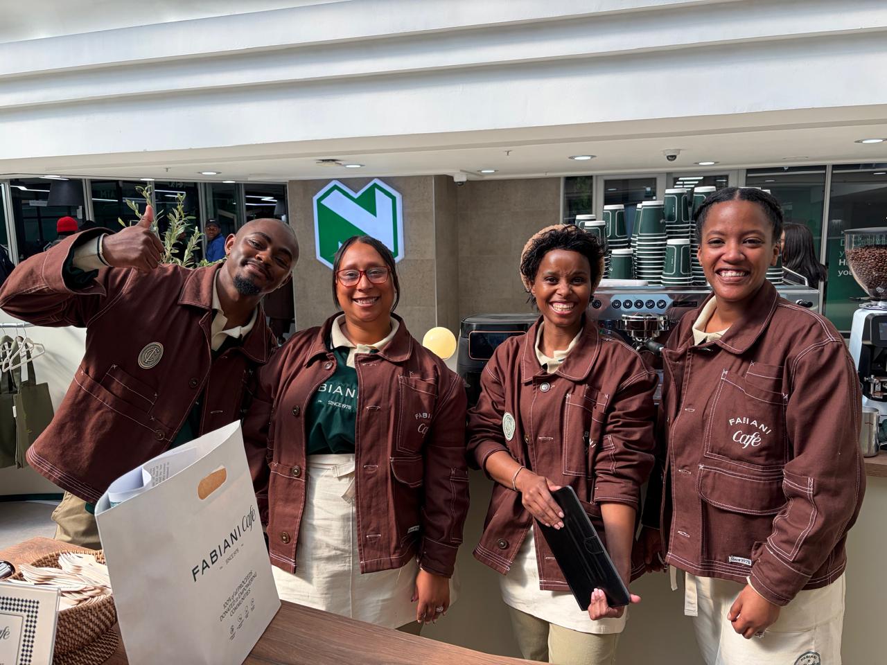 Four smiling SOZO Foundation barista graduates in brown Fabiani Café workwear jackets, posing behind the pop-up counter