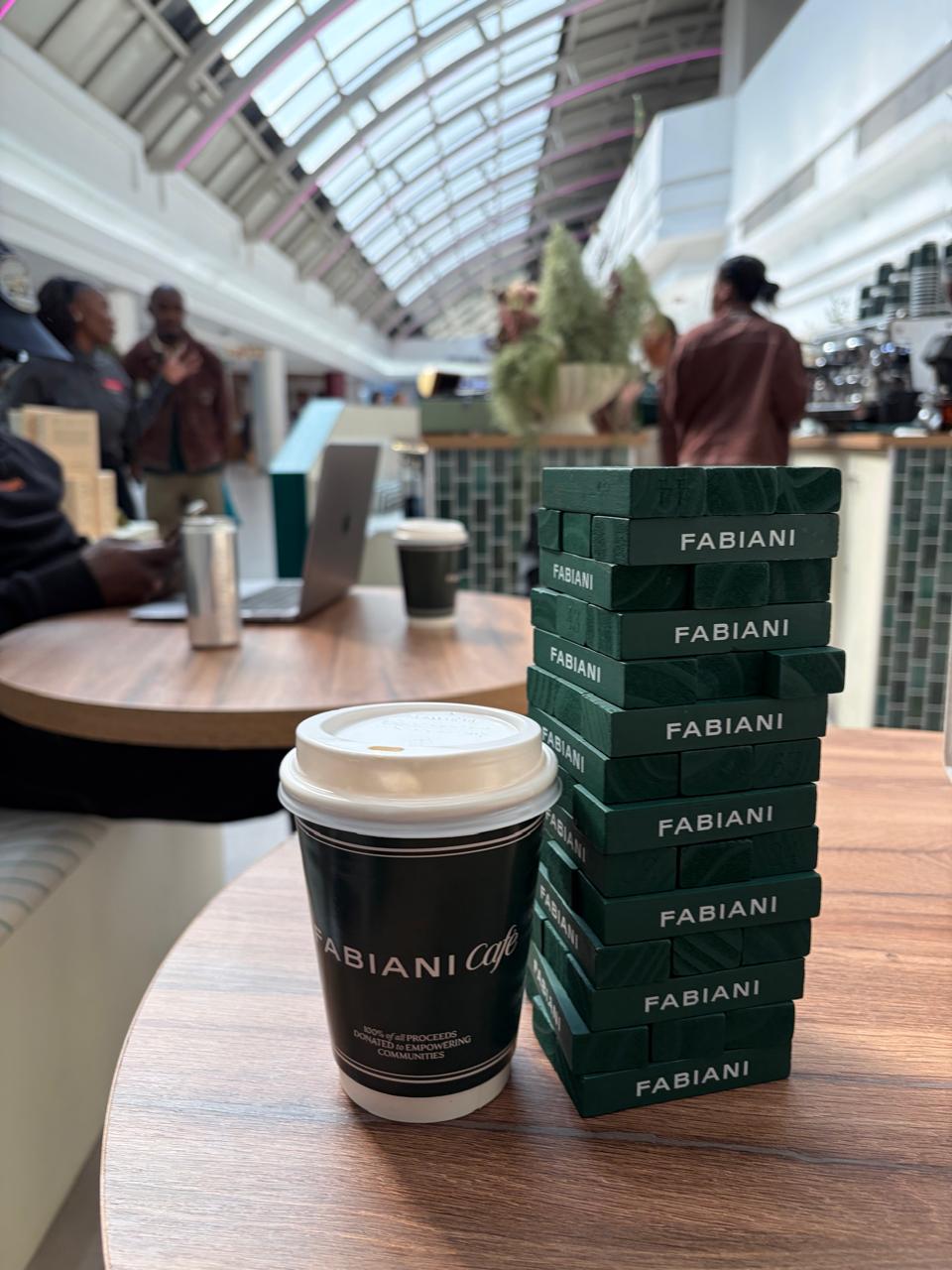 A Fabiani Café takeaway cup next to a stack of green Fabiani-branded Jenga blocks, with the espresso bar and customers visible in the background
