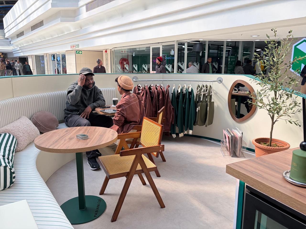 The Fabiani Café lounge area with curved white seating, cane-back chairs, a clothing rail of brown and green jackets, and an olive tree