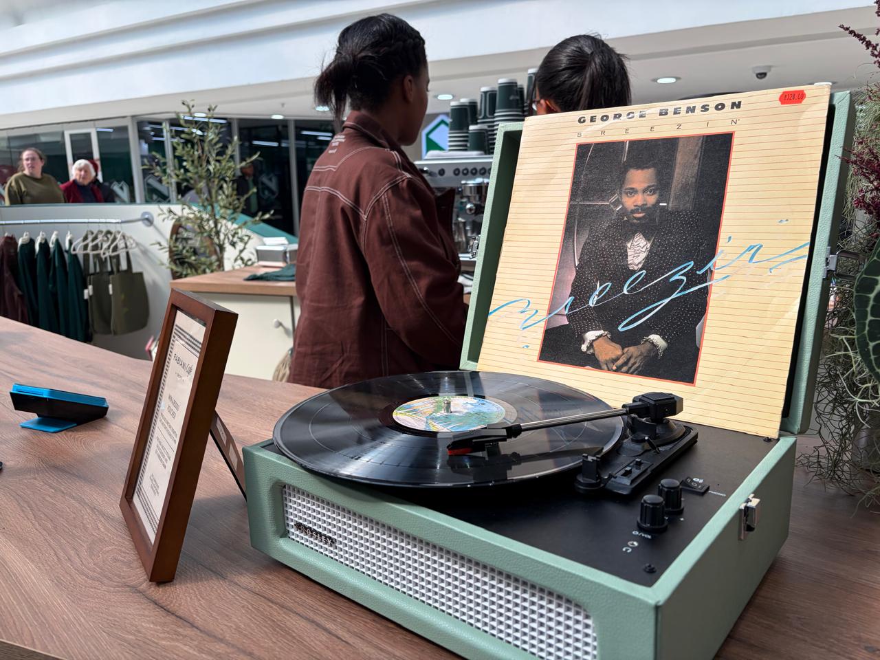 A green vintage-style record player spinning George Benson's Breezin' album at the Fabiani Café, with an olive plant and the barista station in the background