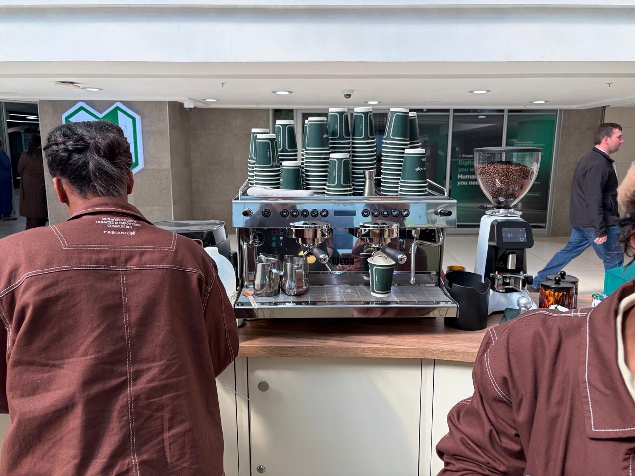 Close-up of the Wega espresso machine behind the Fabiani Café counter, with green cups stacked and a barista at work