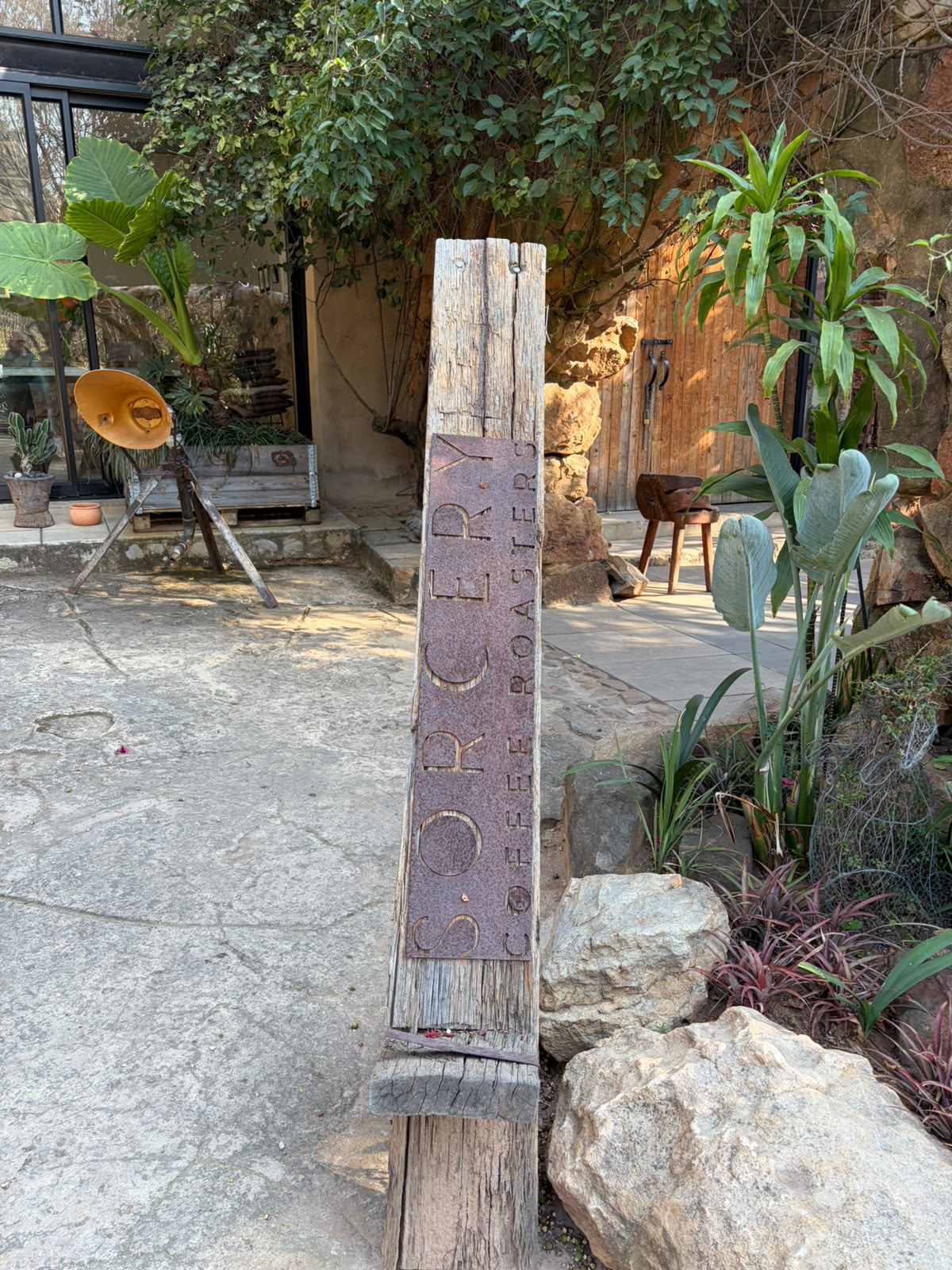 The Sorcery Coffee Roasters entrance sign — a weathered wooden post with hammered copper lettering, nestled among rocks and tropical plants at The Ruins in Wapadrand
