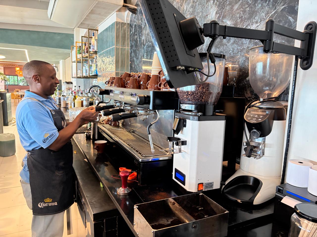 Barista in blue polo and cross-back apron pulling a shot on a CPEM espresso machine, brown cups stacked on top, dark marble wall behind