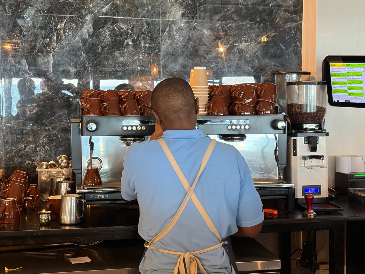 Barista working the CPEM machine from a wider angle — grinder on the right, coffee jiggers and steel jugs on the left, POS system visible