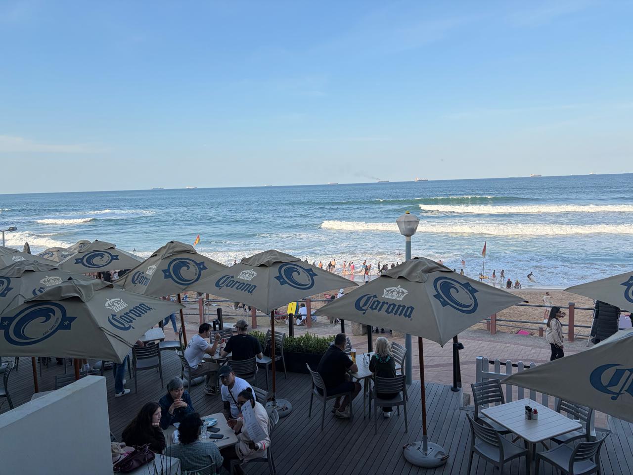 Outdoor terrace at Surf Riders — Corona umbrellas, beachgoers, Indian Ocean waves, ships on the horizon