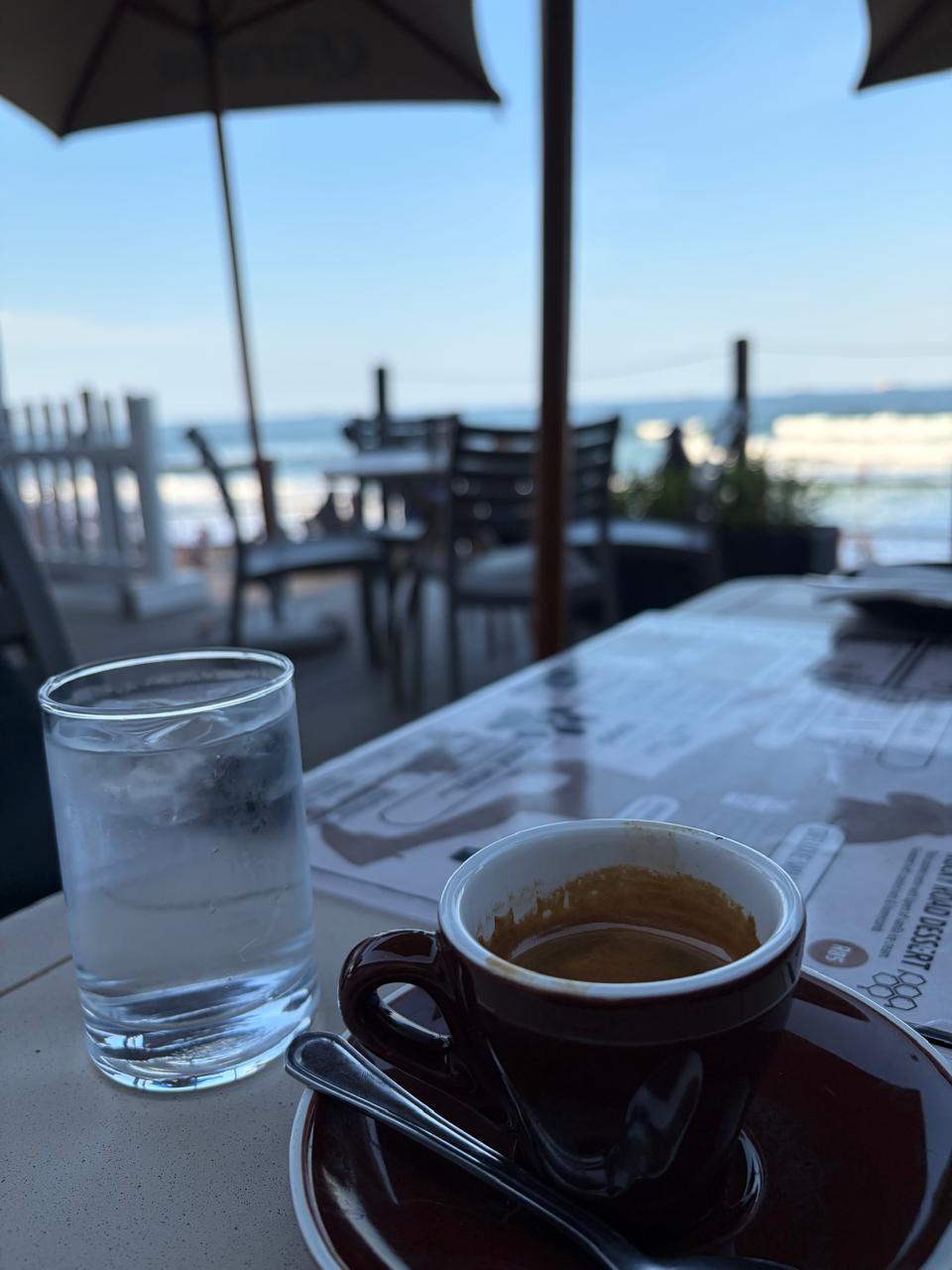 Espresso in a brown cup with water glass at Surf Riders — ocean waves and beach umbrellas visible in the background