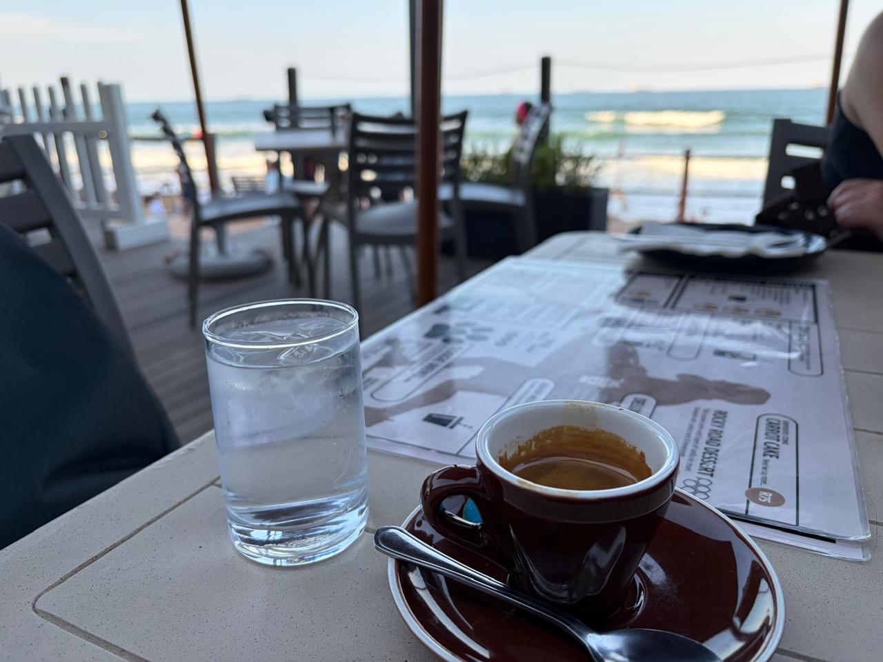 Another angle of the espresso — brown cup, glass of water, ocean and deck chairs visible behind