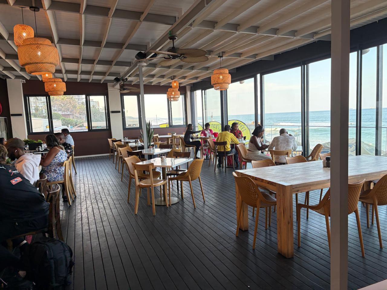 Wide-angle interior of Surf Riders Café — wooden tables, rattan pendant lights, floor-to-ceiling glass with ocean views, dark timber deck floors