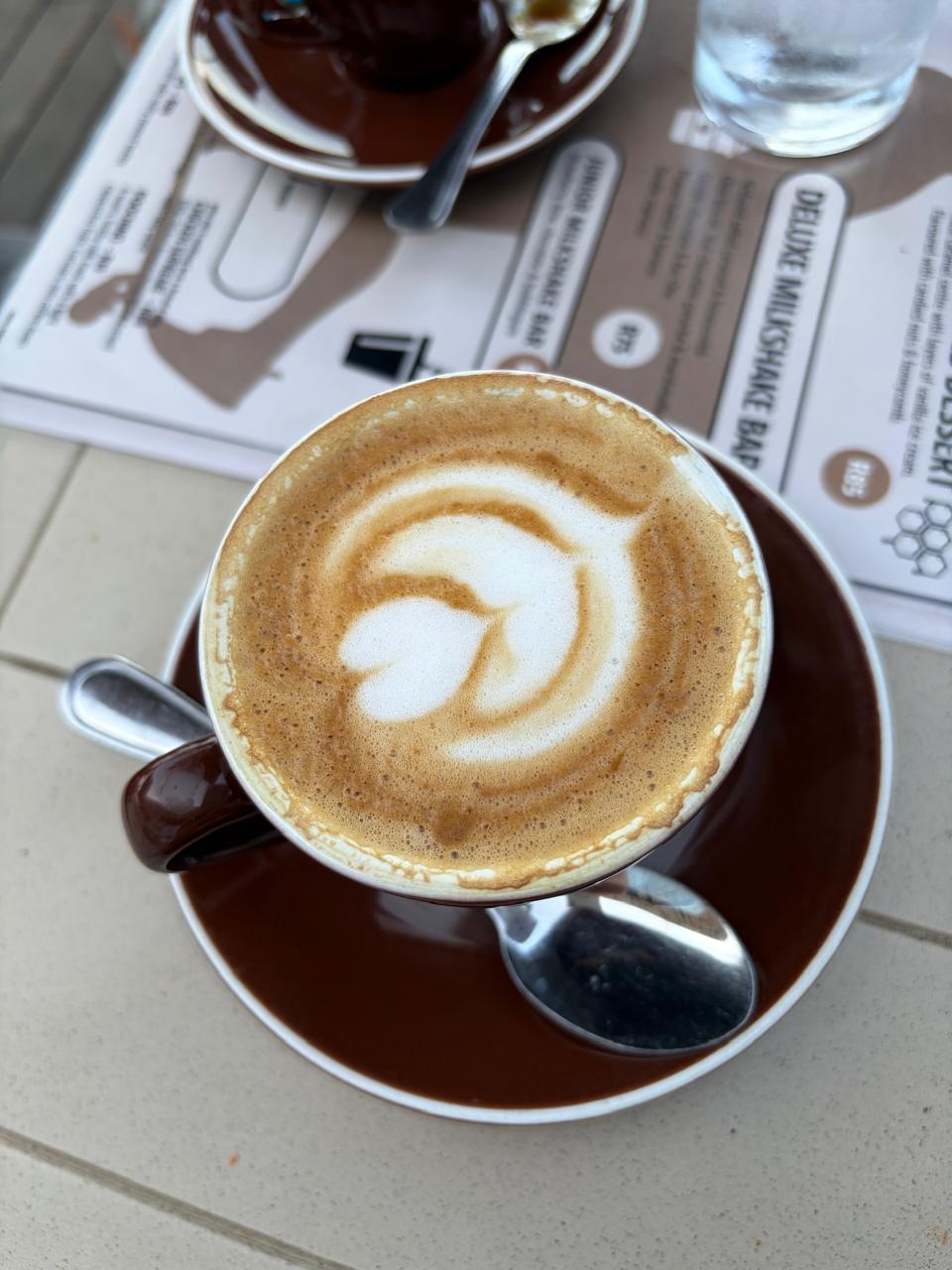 Top-down view of a flat white with heart latte art in a brown cup on a brown saucer, table setting visible
