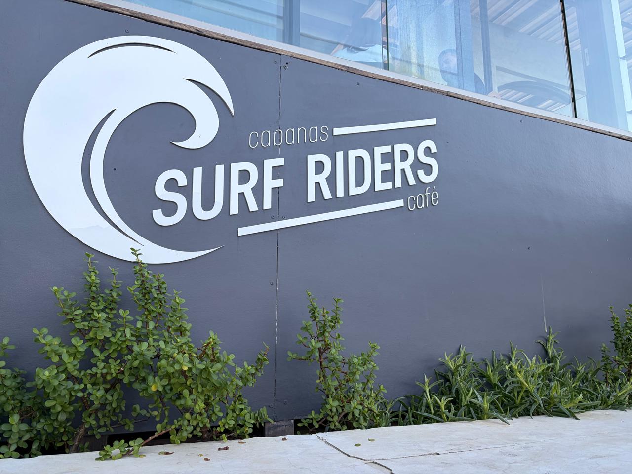 Cabanas Surf Riders Café exterior sign — wave logo, dark grey wall, green succulents in the landscaping