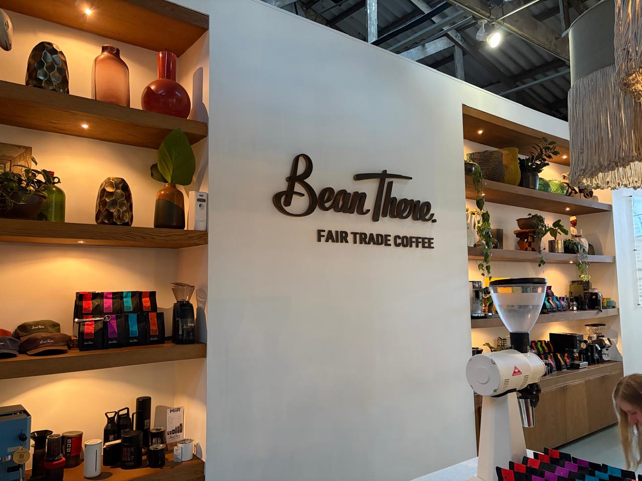 Bean There Fair Trade Coffee logo on the white wall of the Milpark café, with retail shelves of colourful bean bags and a Mahlkönig grinder in the foreground