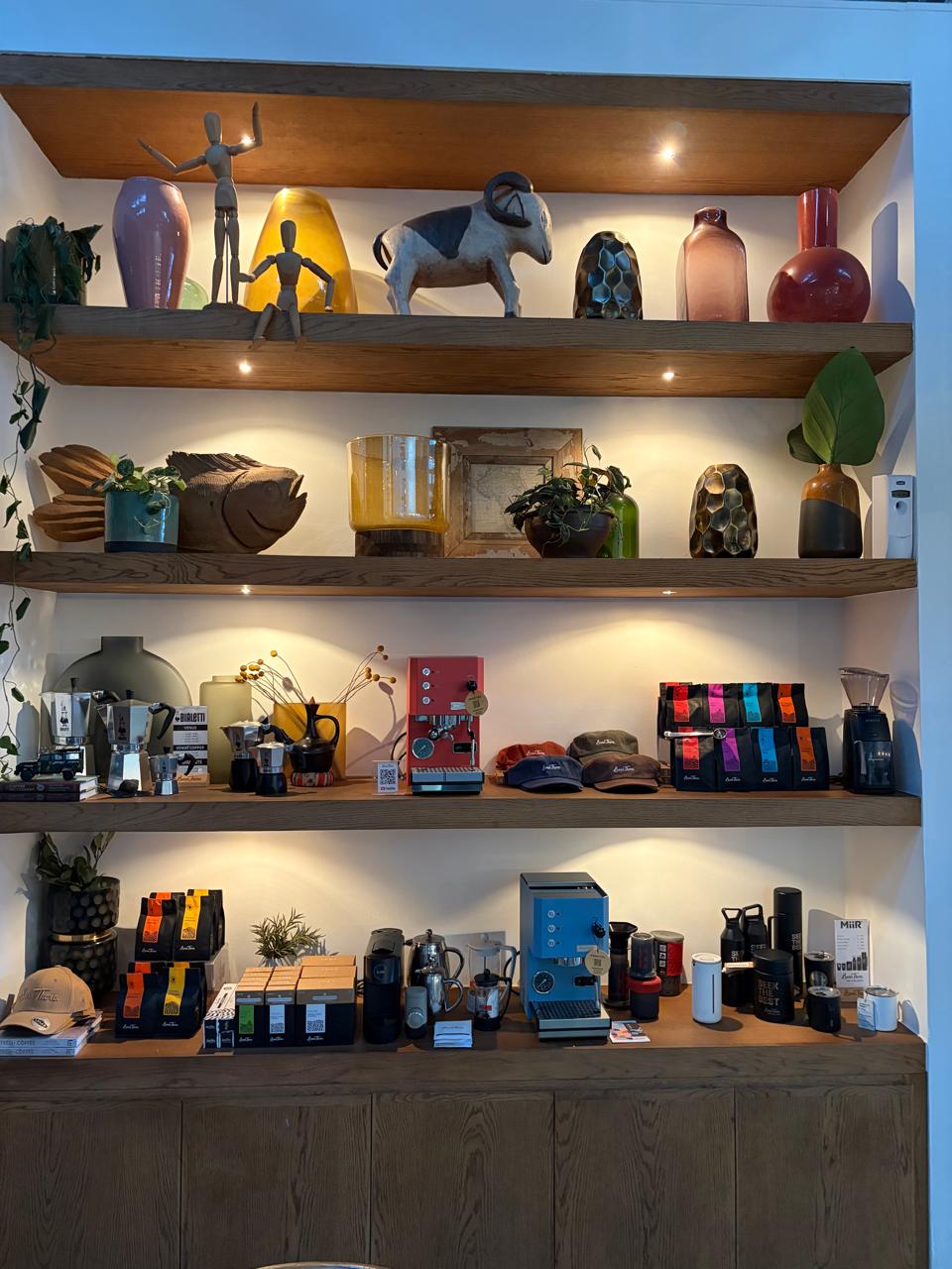 Bean There retail shelves — Bialetti moka pots, espresso machines in red and blue, MiiR tumblers, Bean There caps, and bags of single-origin coffee from every African partner country
