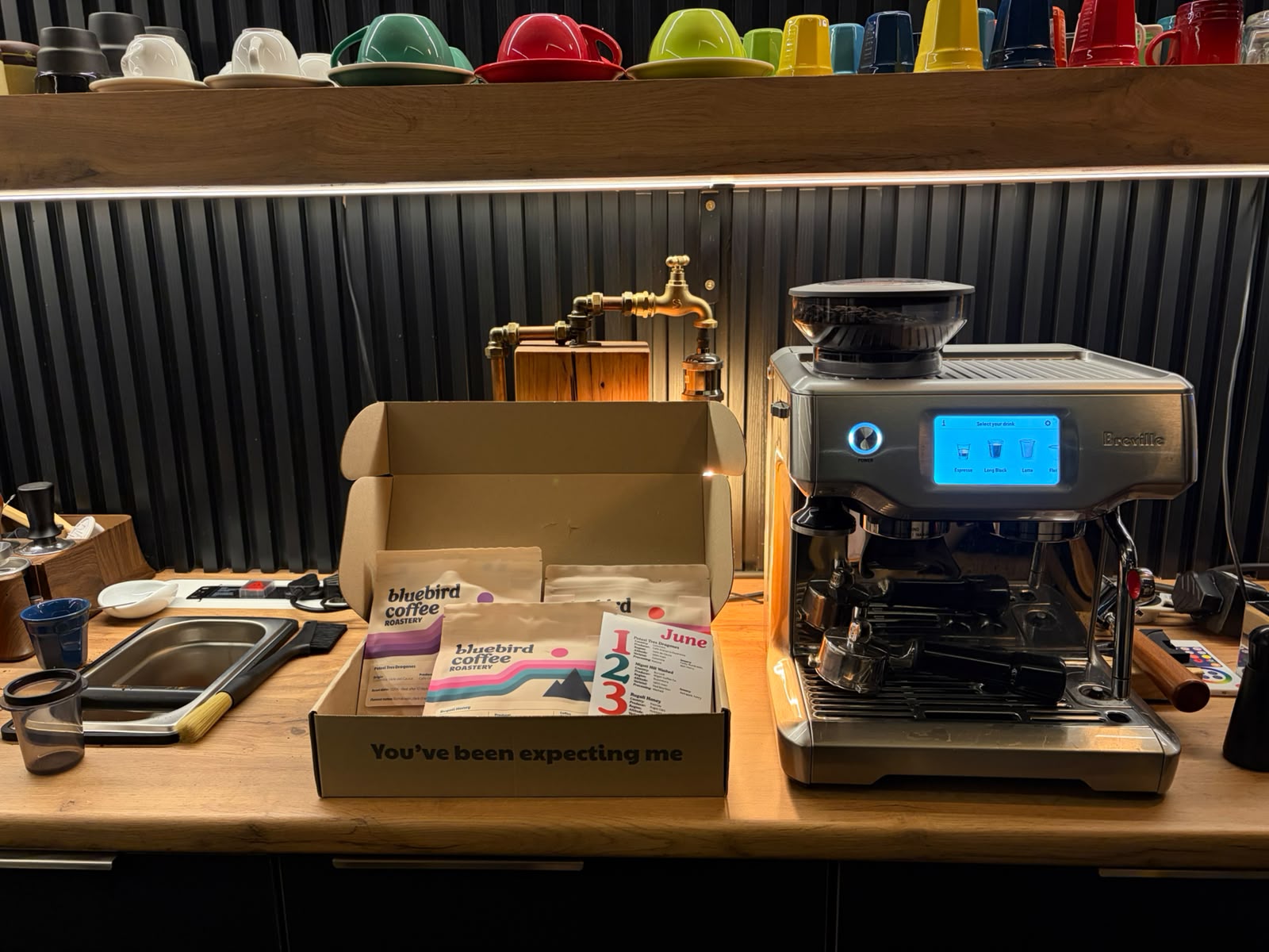 Full home espresso setup with Bluebird Coffee Roastery subscription box, Breville machine, and colourful mugs