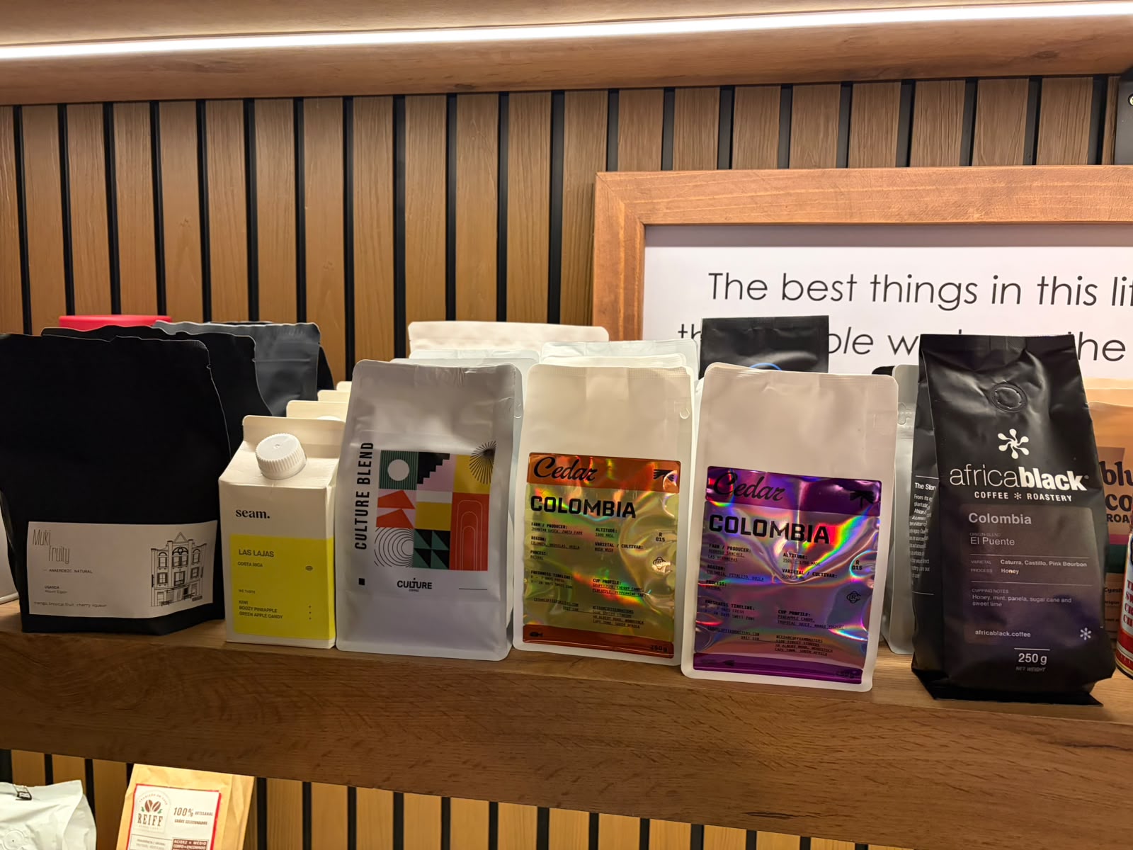 Cedar Colombia holographic bags on a wooden shelf alongside Seam Las Lajas, Culture Blend, and AfricaBlack Colombia El Puente, with a framed quote in the background